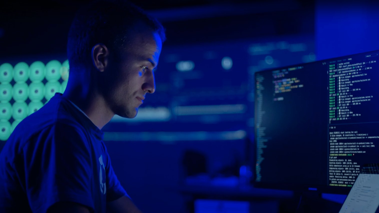 Kent sitting in a dark server room in front of a computer with code up and running