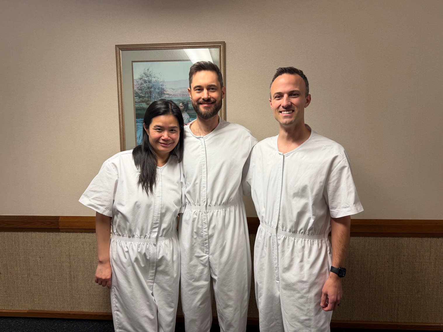 Beiyi, Jeremiah Dow, and Kent C. Dodds stand side by side in white baptismal clothing, smiling indoors at a church.