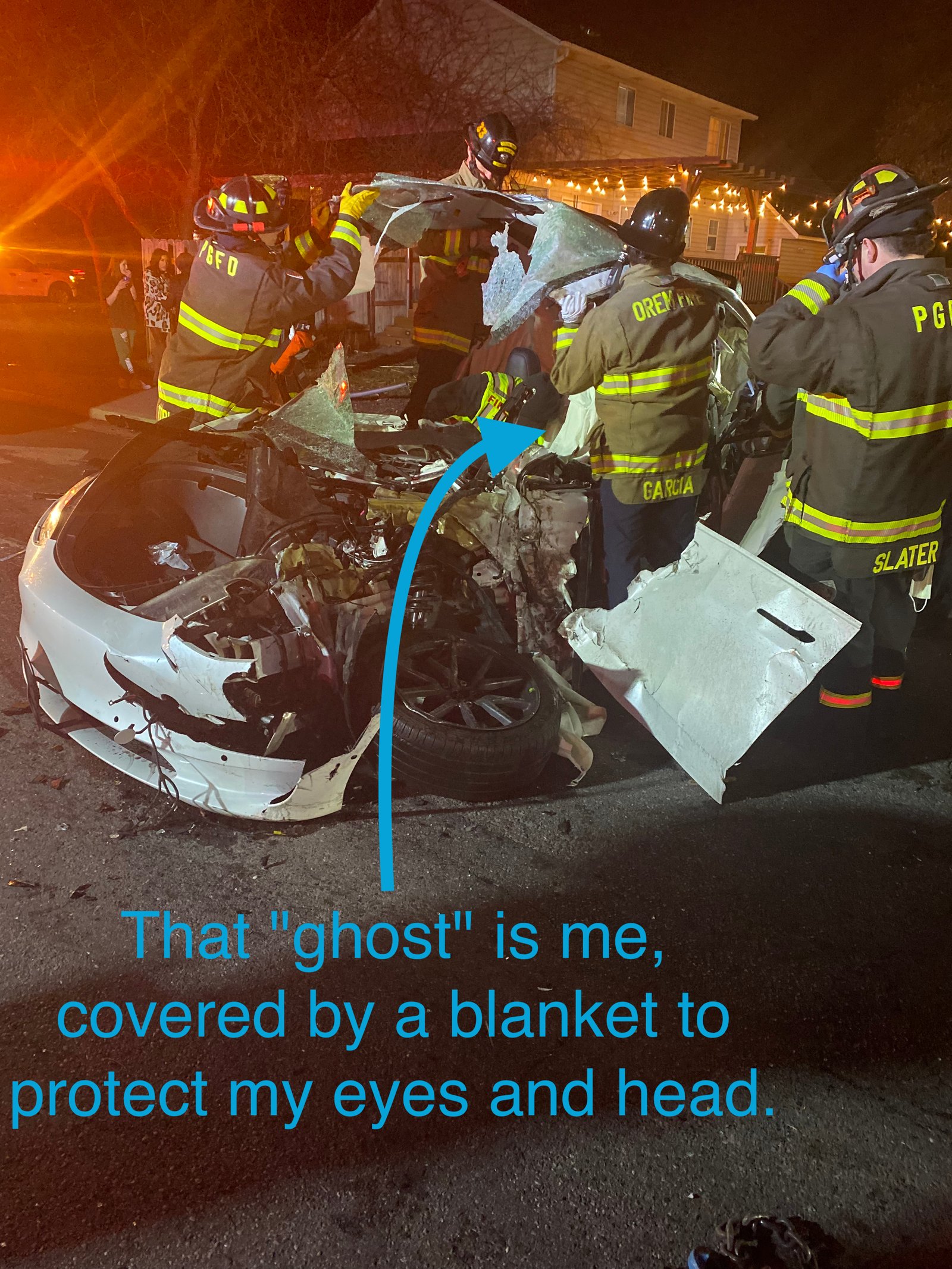 A destroyed Tesla model 3 with firefighters around helping to extract Kent who is covered in a white sheet. An arrow is pointed at Kent with the words: That "ghost" is me, covered by a blanket to protect my eyes and head.